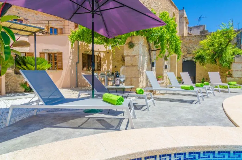 Una terraza exterior con piscina tiene asientos reclinables, una sombrilla morada y vegetación.