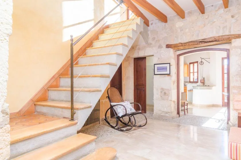 Interior de paredes de piedra con una escalera, una mecedora y una entrada a una habitación luminosa.