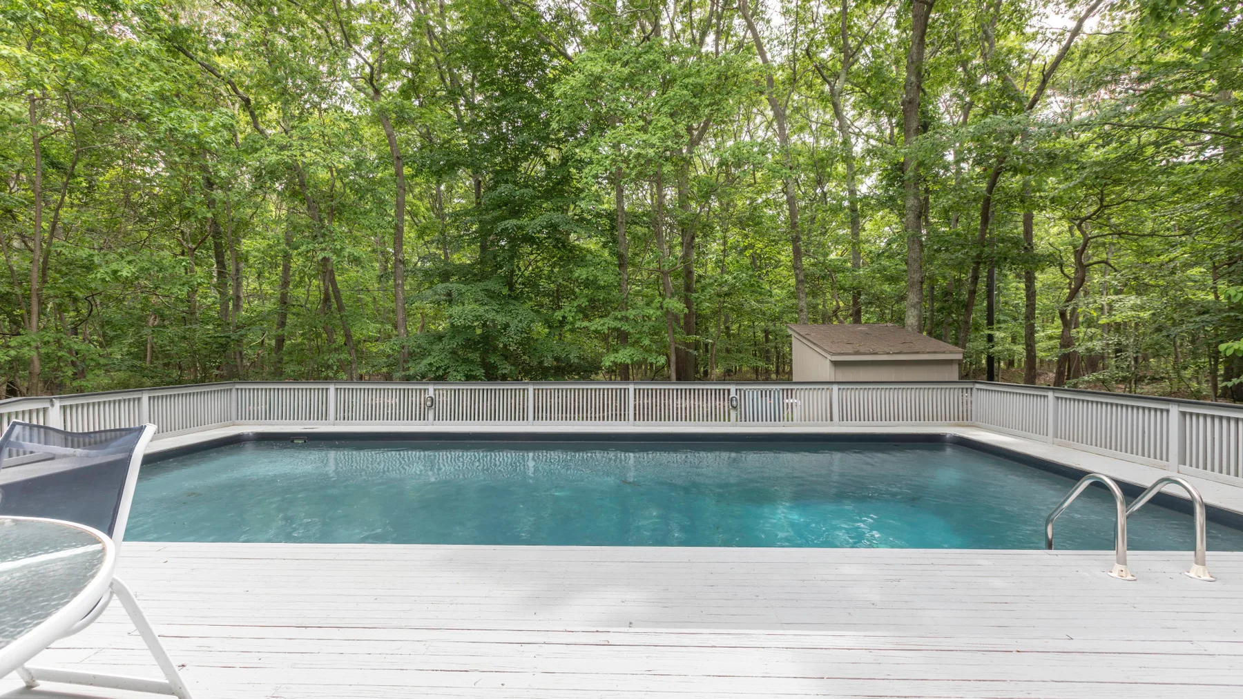 Outdoor swimming pool beside a wooded area, with a broad deck.