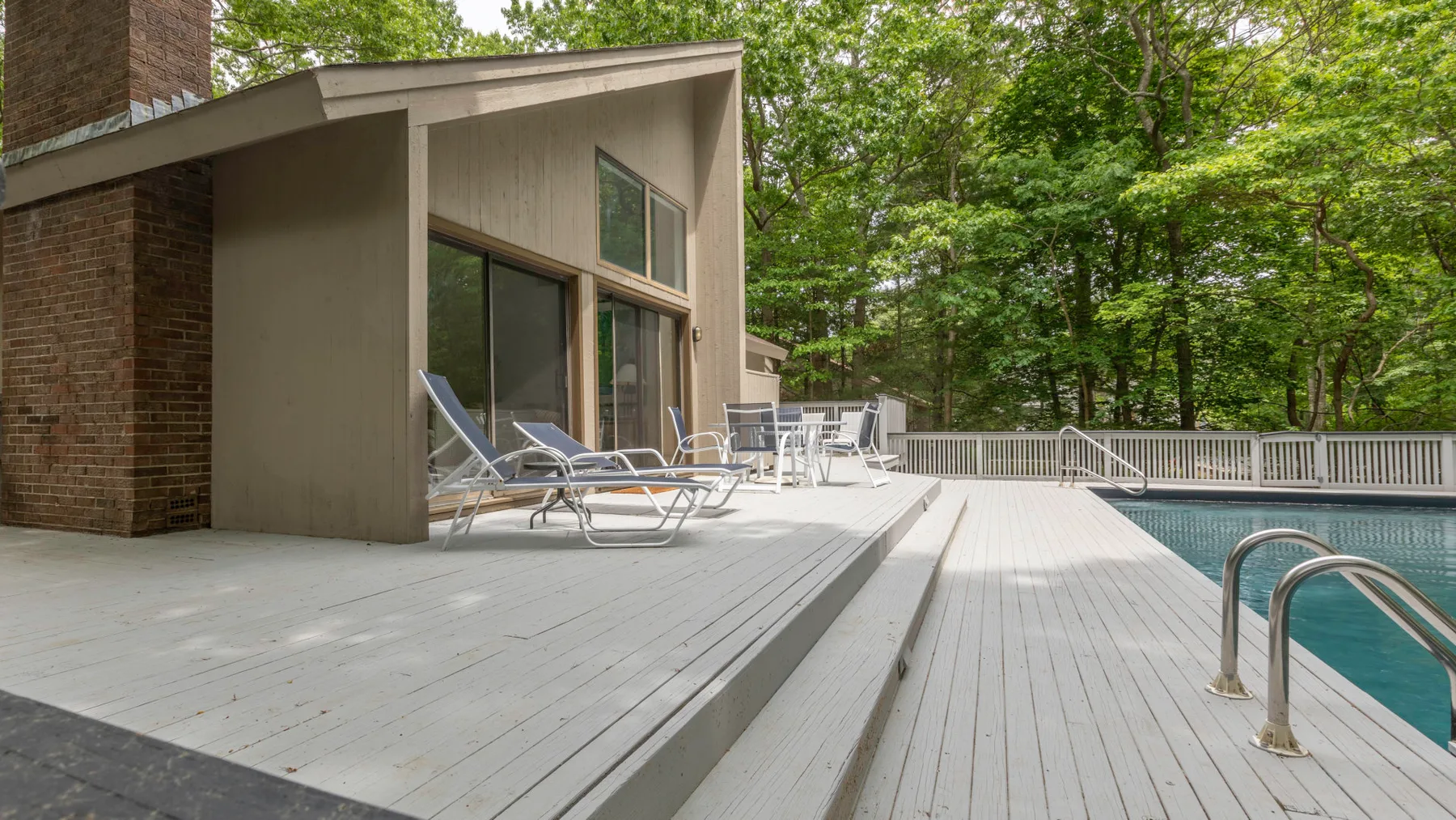 A poolside deck extends beside a modern building, with trees in the background.