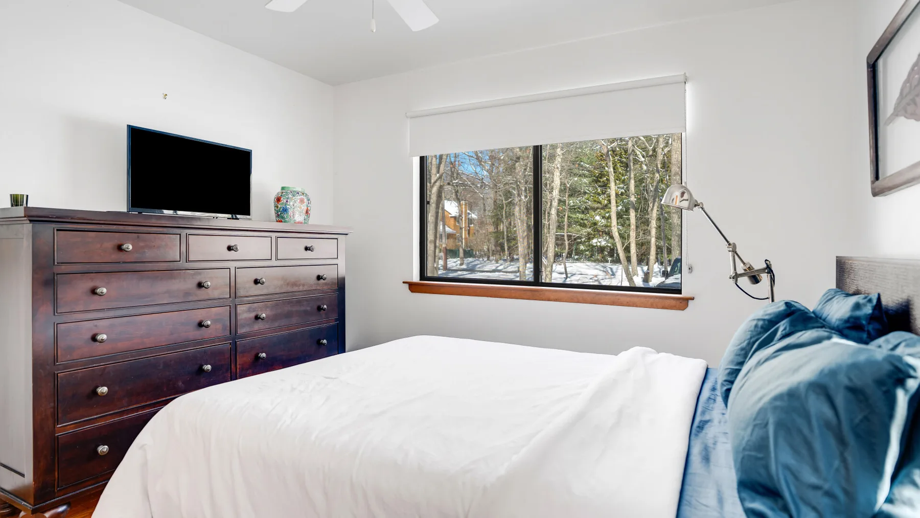 A bright bedroom with a bed, dresser, and large window overlooking trees.