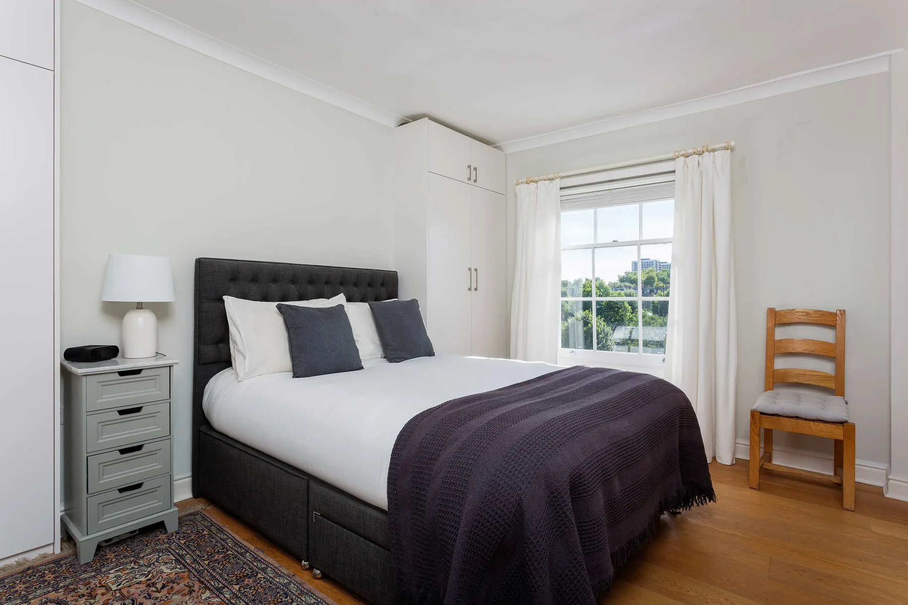 A bedroom with a bed, bedside table, and a window with white curtains.