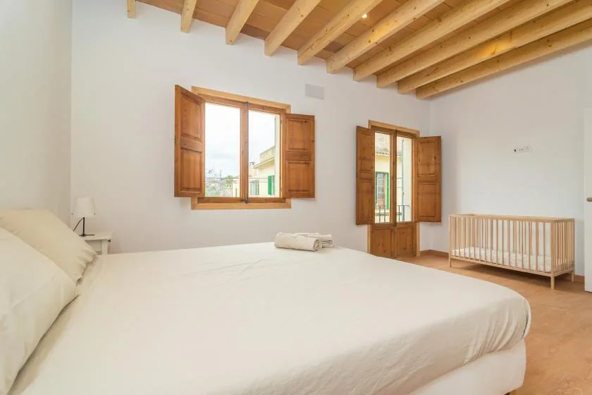 Un dormitorio luminoso con una cama grande, vigas de madera y contraventanas abiertas.