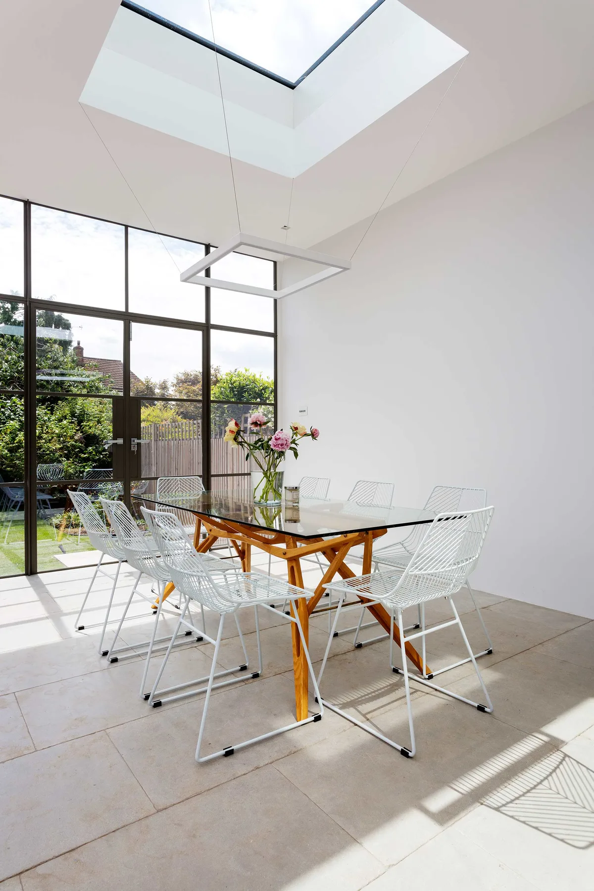 Un comedor luminoso con una mesa de vidrio y asientos blancos de alambre junto a grandes ventanas.