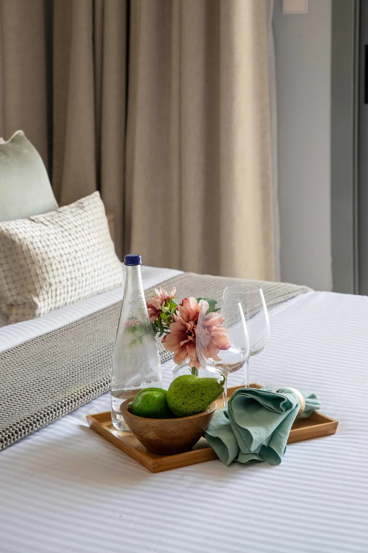 Una cama bien arreglada con cortinas neutras y una bandeja con fruta y flores.