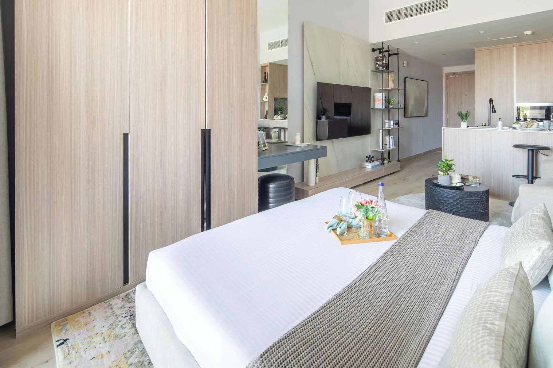 Dormitorio moderno con una cama bien tendida y una pequeña cocina al fondo.