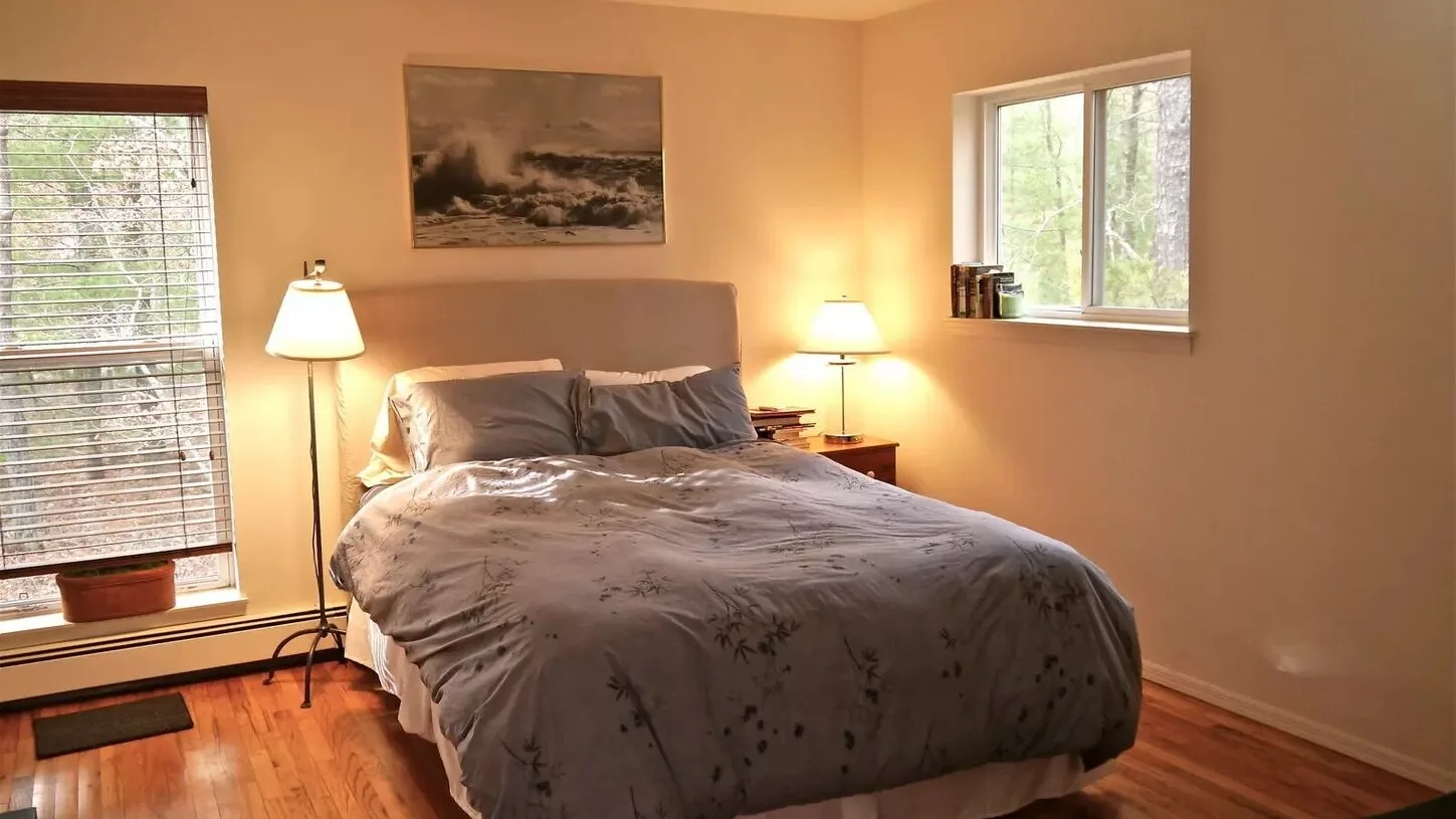 Un dormitorio con una cama, mesitas de noche y ventanas por donde entra la luz.
