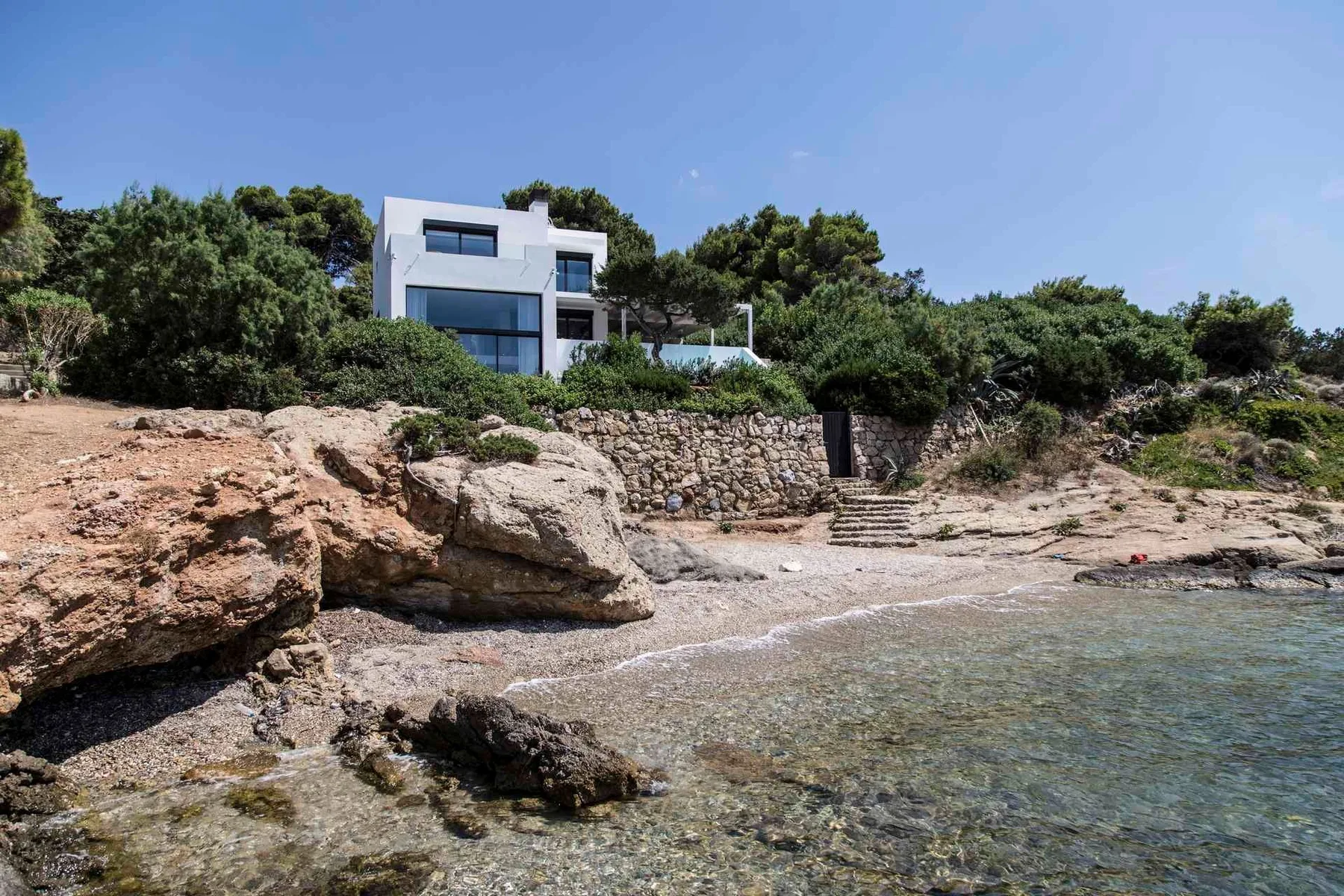 Una casa blanca moderna se alza sobre una costa rocosa junto al agua clara.