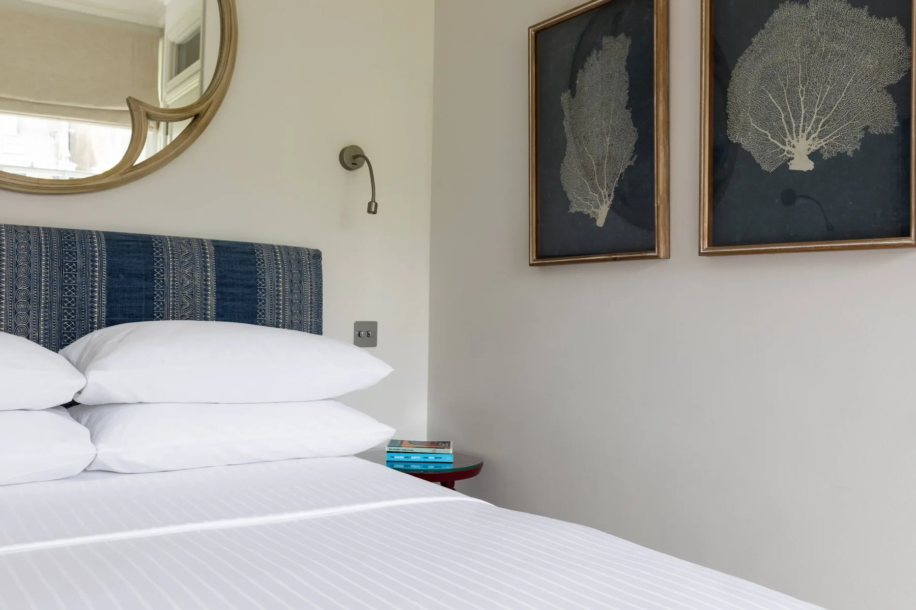A bedroom with a neatly made bed, framed botanical prints, and a round mirror.