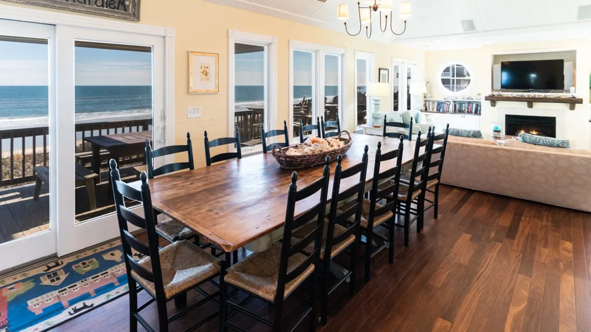 Comedor con una larga mesa de madera junto a grandes ventanas con vistas al agua.
