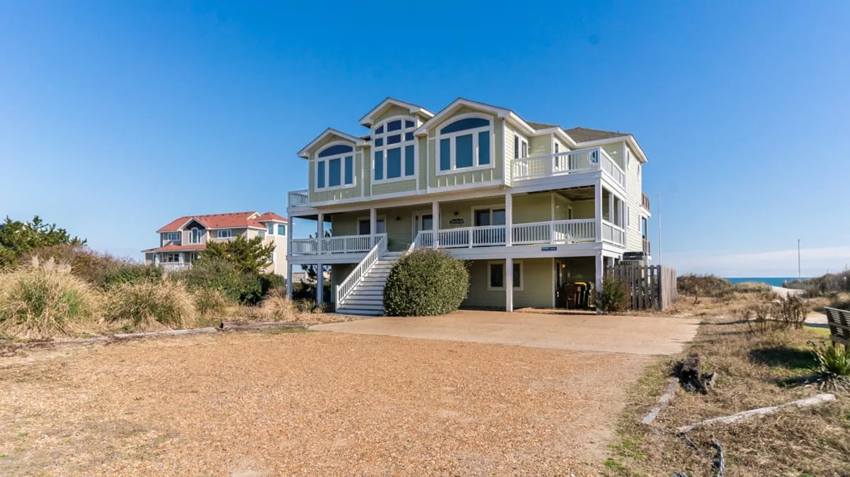 Una casa costera grande con balcones elevados se alza junto a un patio de arena.