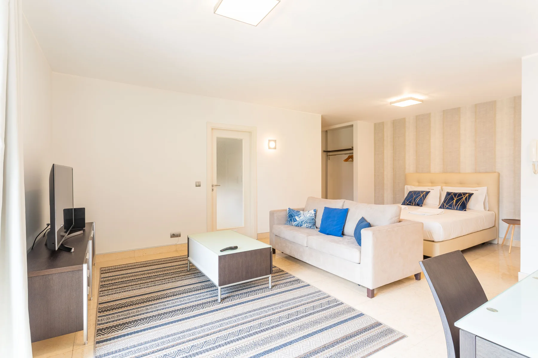 A bright studio room combines a bed, seating area, and striped rug.