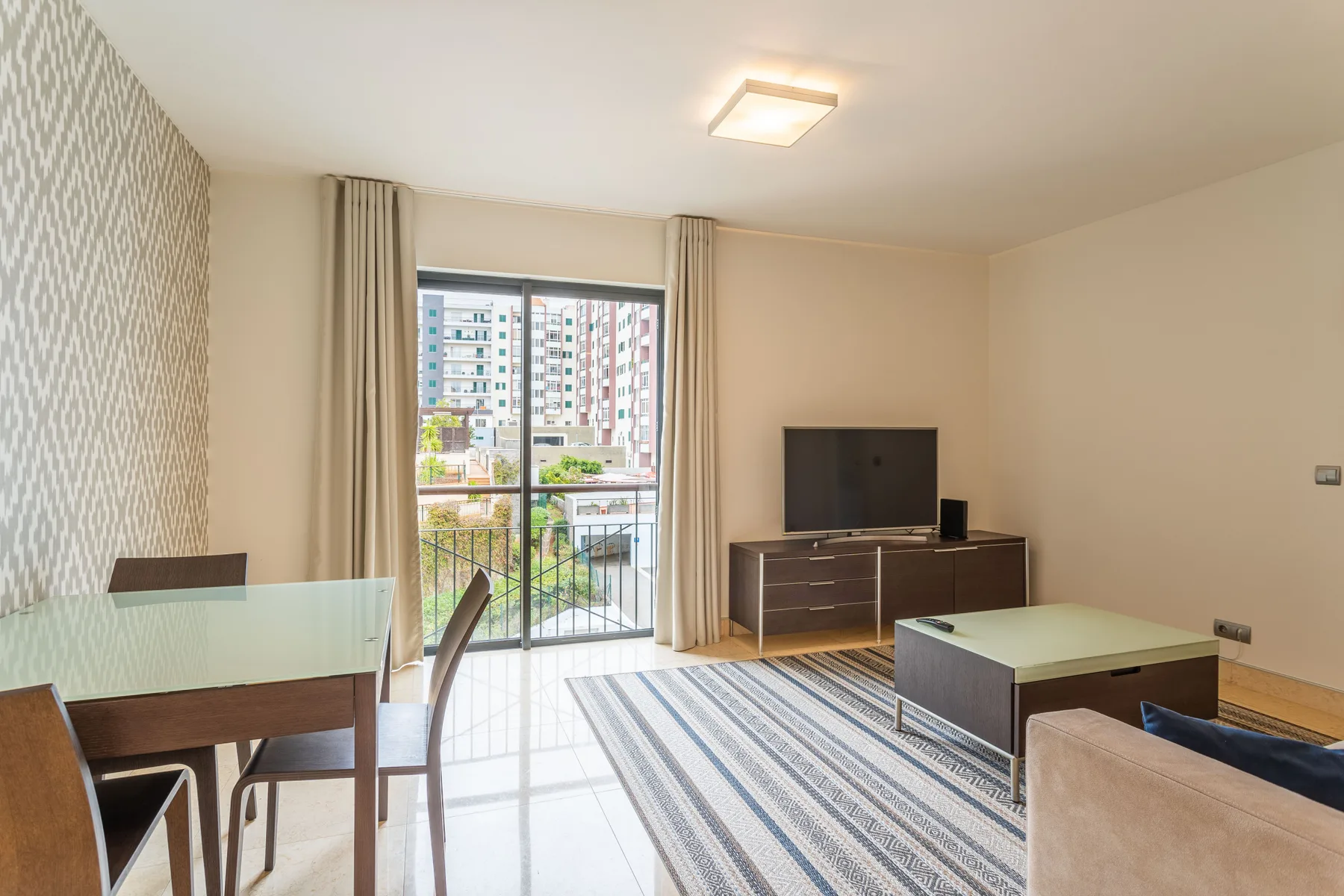 A bright studio living area with a dining table, television, and balcony doors.
