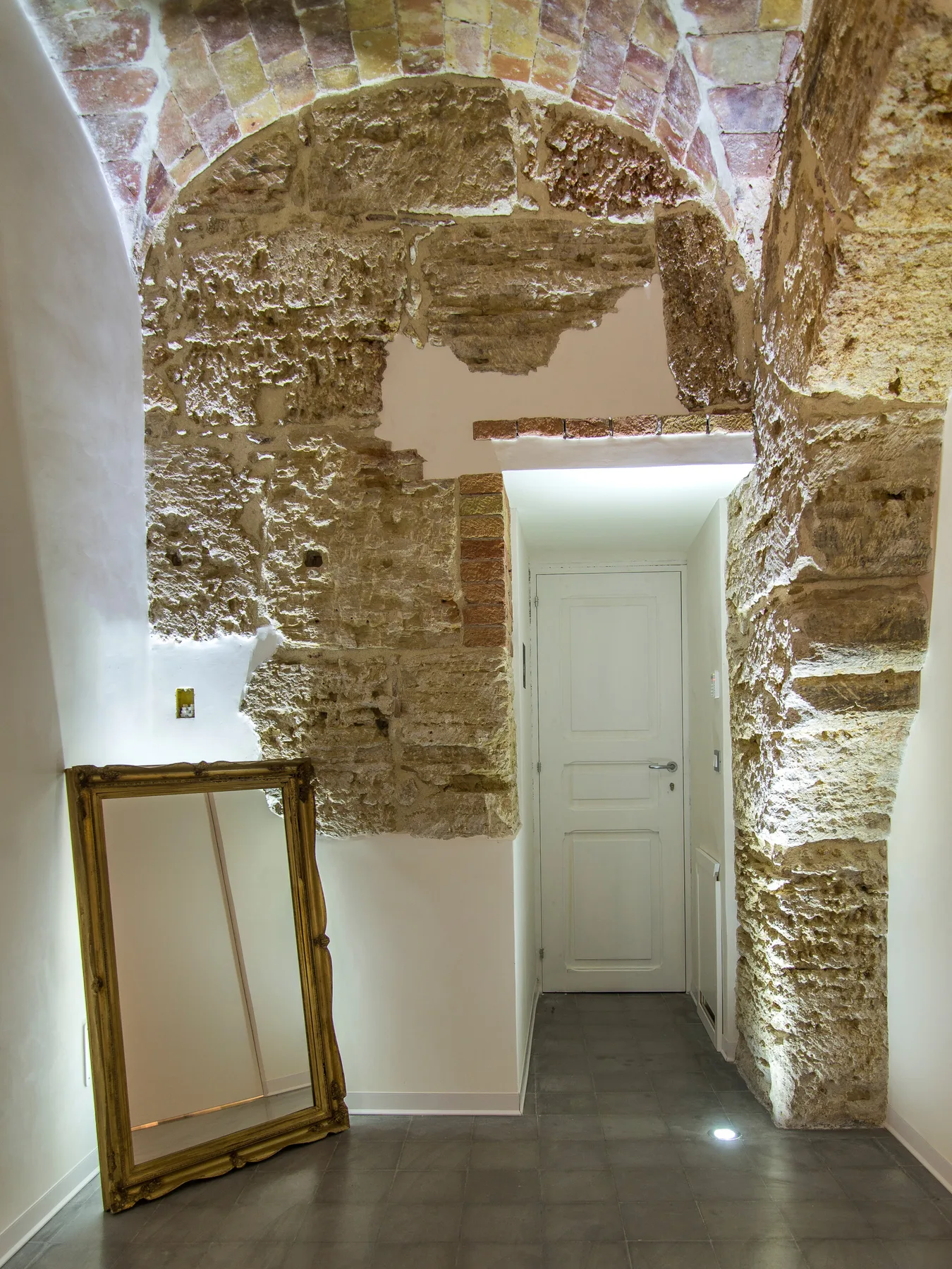 A stone arched hallway with a white door and a framed mirror leaning against the wall.