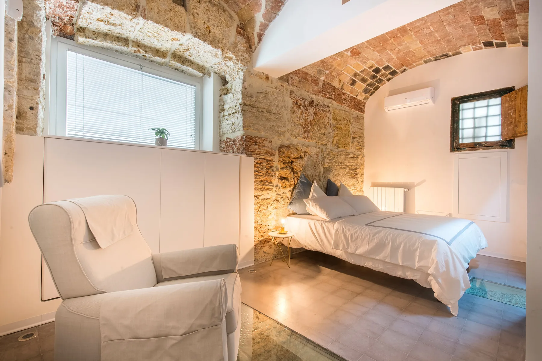 A bedroom with a bed and armchair beneath an exposed stone arch.