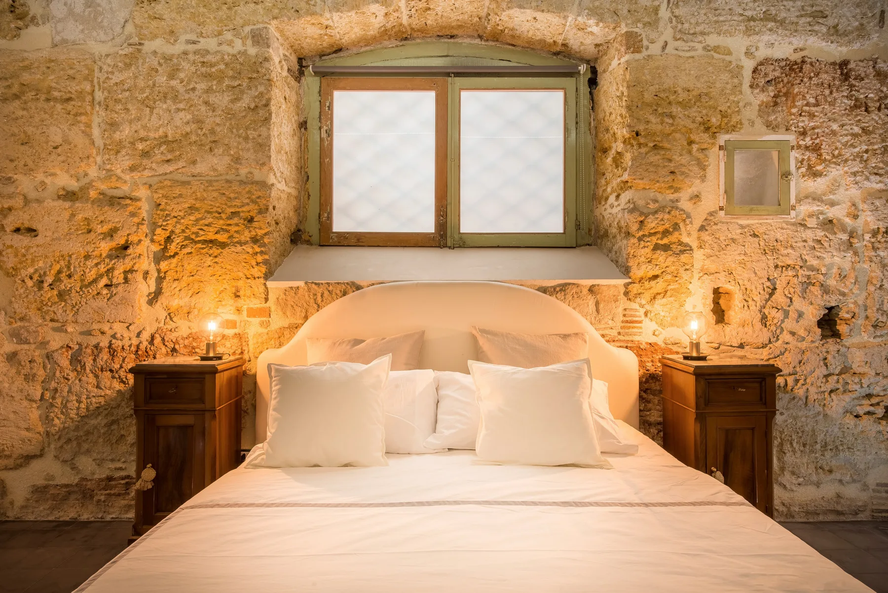 A bedroom with a bed beneath a small window set in a stone wall.