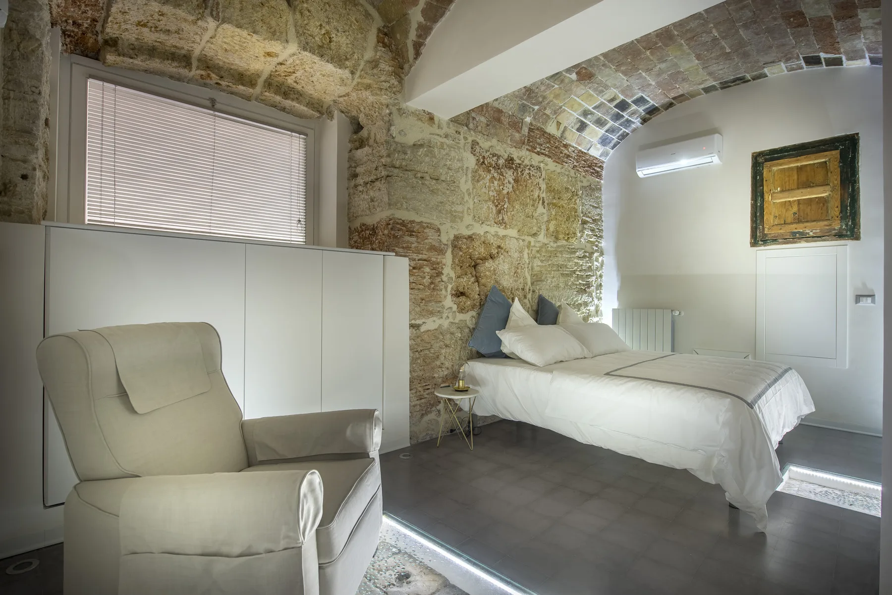 A bedroom with a bed beneath a brick arch and a cushioned armchair.