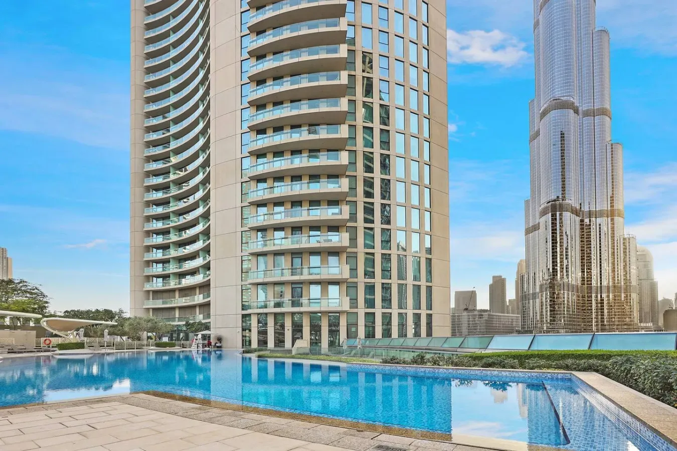 A rooftop swimming pool sits beside modern high-rise buildings under a blue sky.