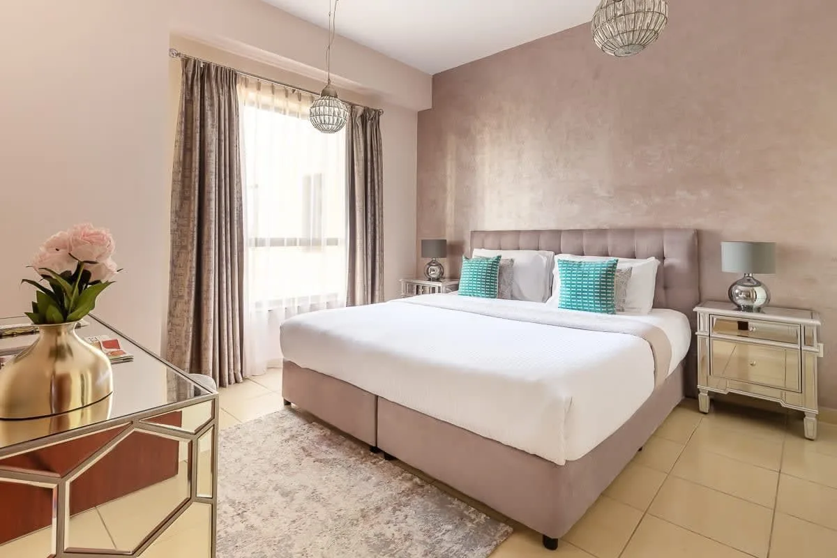 Un dormitorio con una cama bien hecha, cojines turquesa y una gran ventana con cortinas.