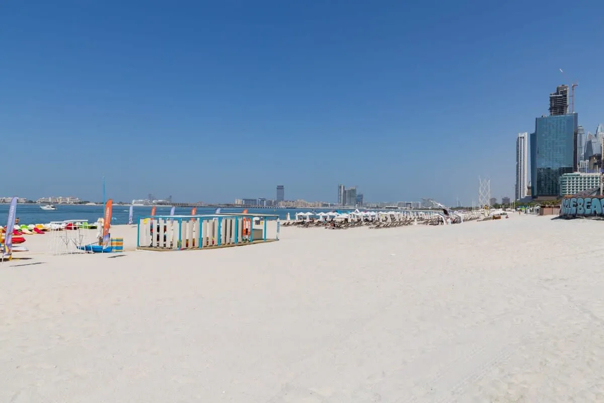 Una amplia playa de arena se extiende bajo un cielo azul despejado, con edificios de la ciudad a lo lejos.