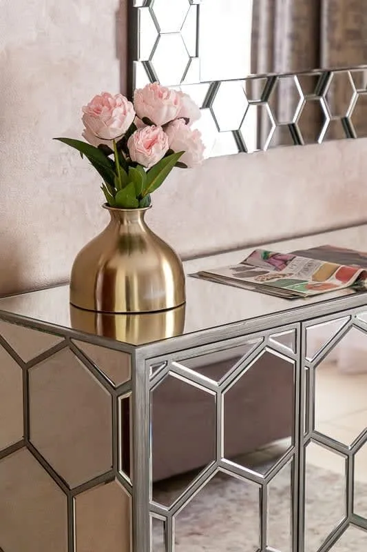 Una consola con espejos sostiene un jarrón dorado con flores rosas bajo un espejo geométrico.