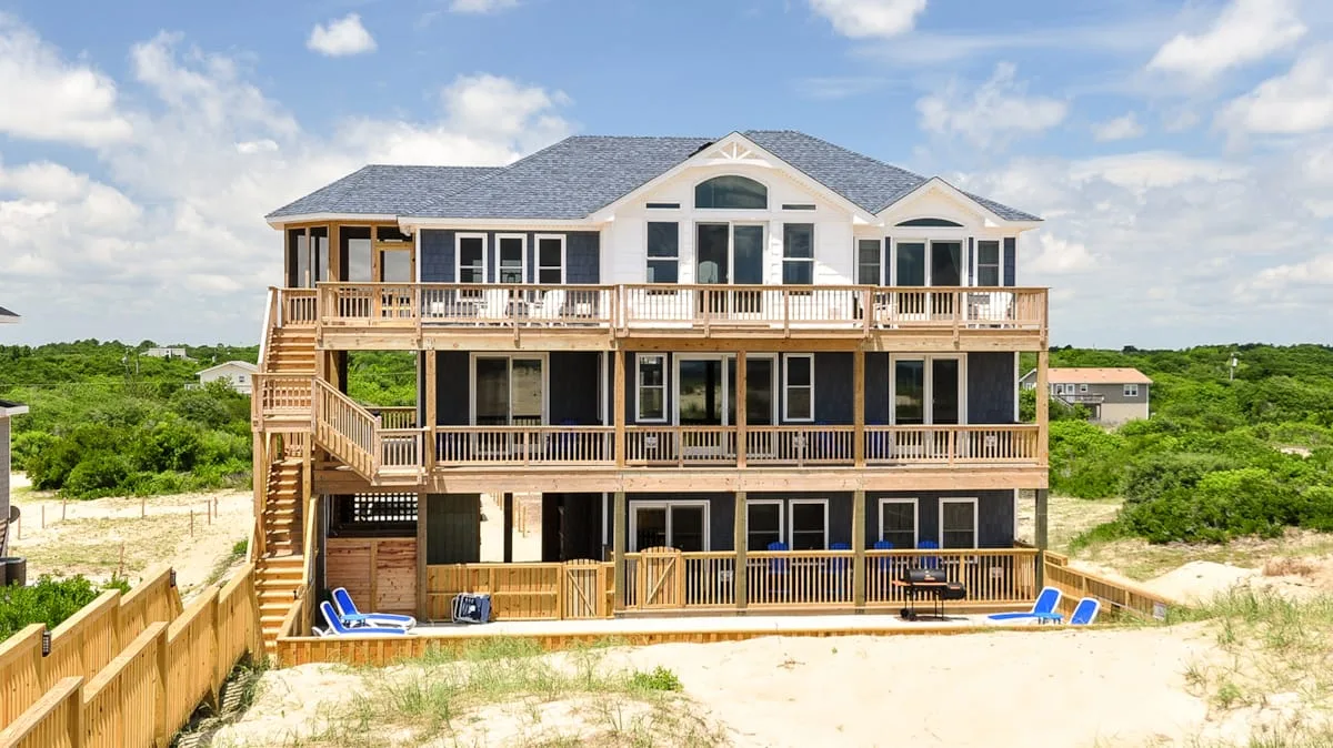 A large coastal house with wooden decks and surrounding sand dunes.