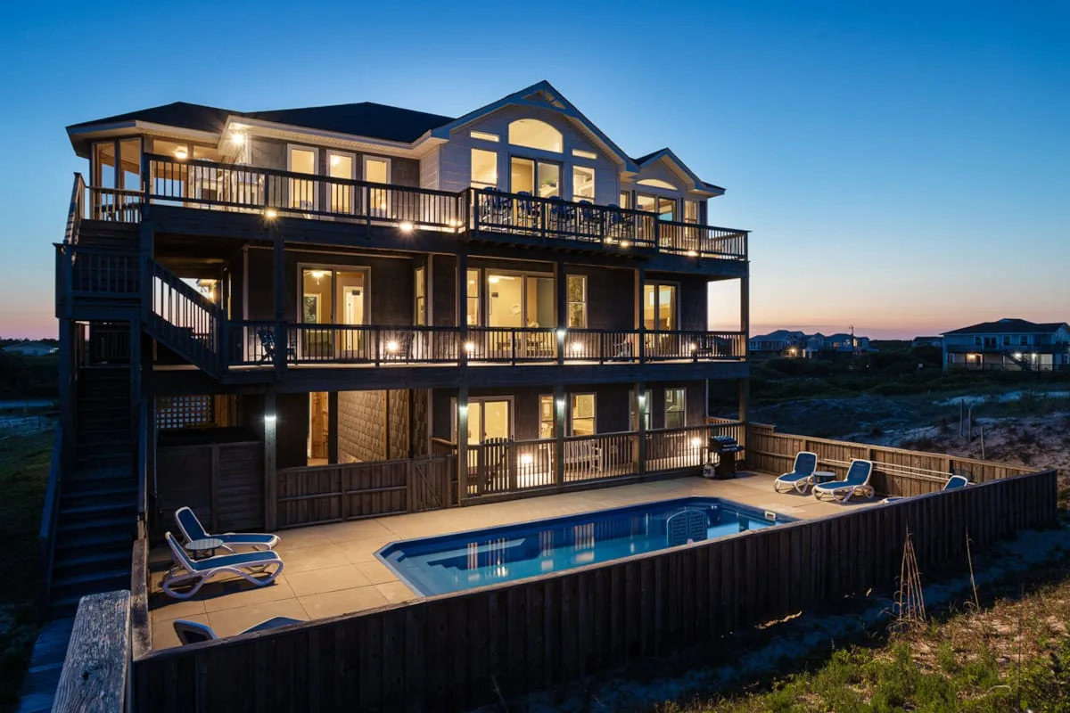 A large house with balconies overlooks an outdoor pool at dusk.