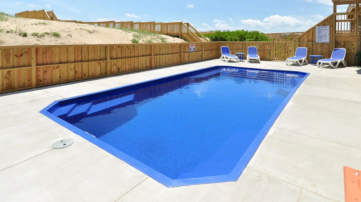 An outdoor swimming pool sits in a paved area beside wooden fencing.