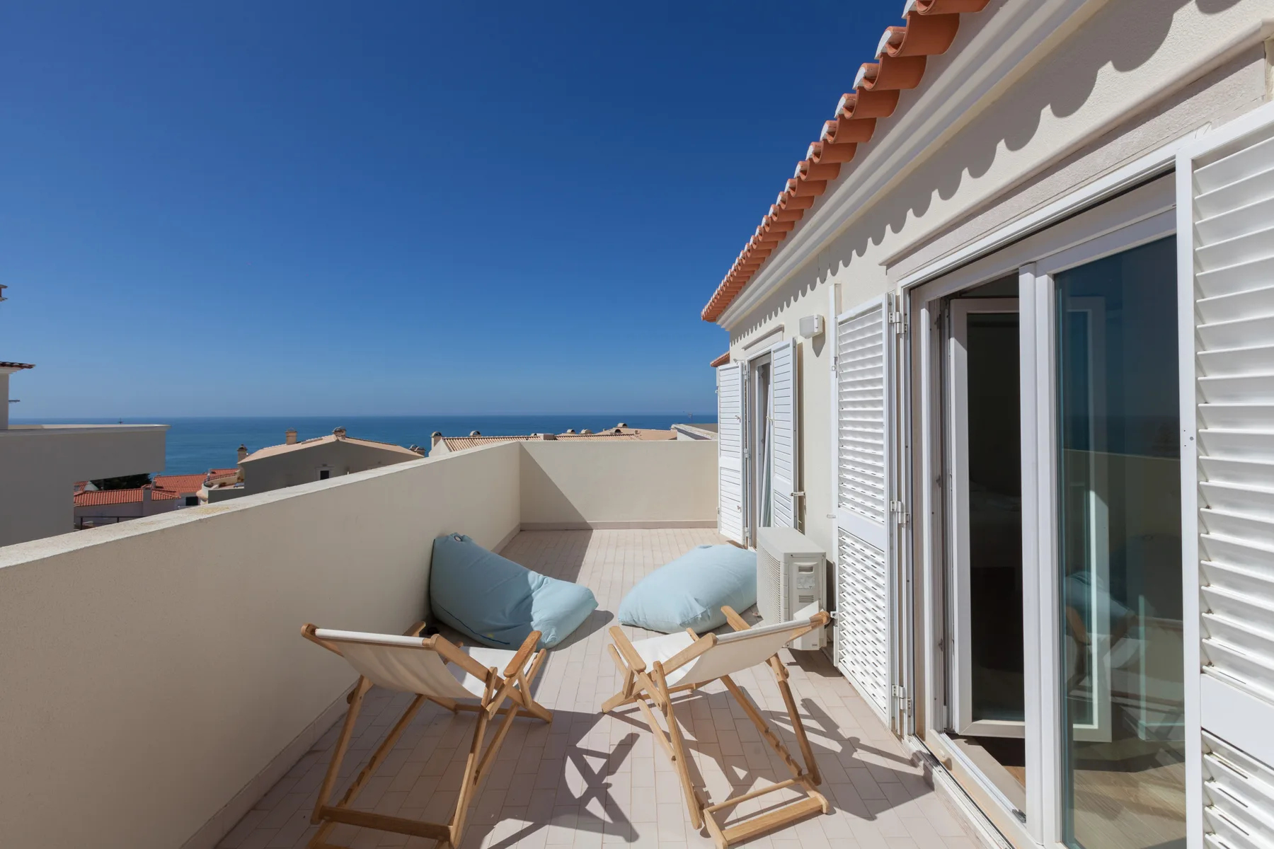 A terrace with folding chairs and cushions overlooks the sea beside a white building.