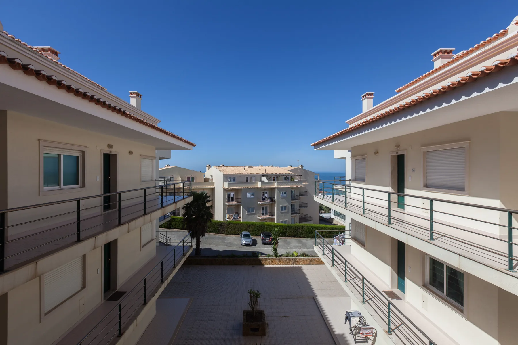A courtyard between apartment buildings overlooks the sea under a clear blue sky.