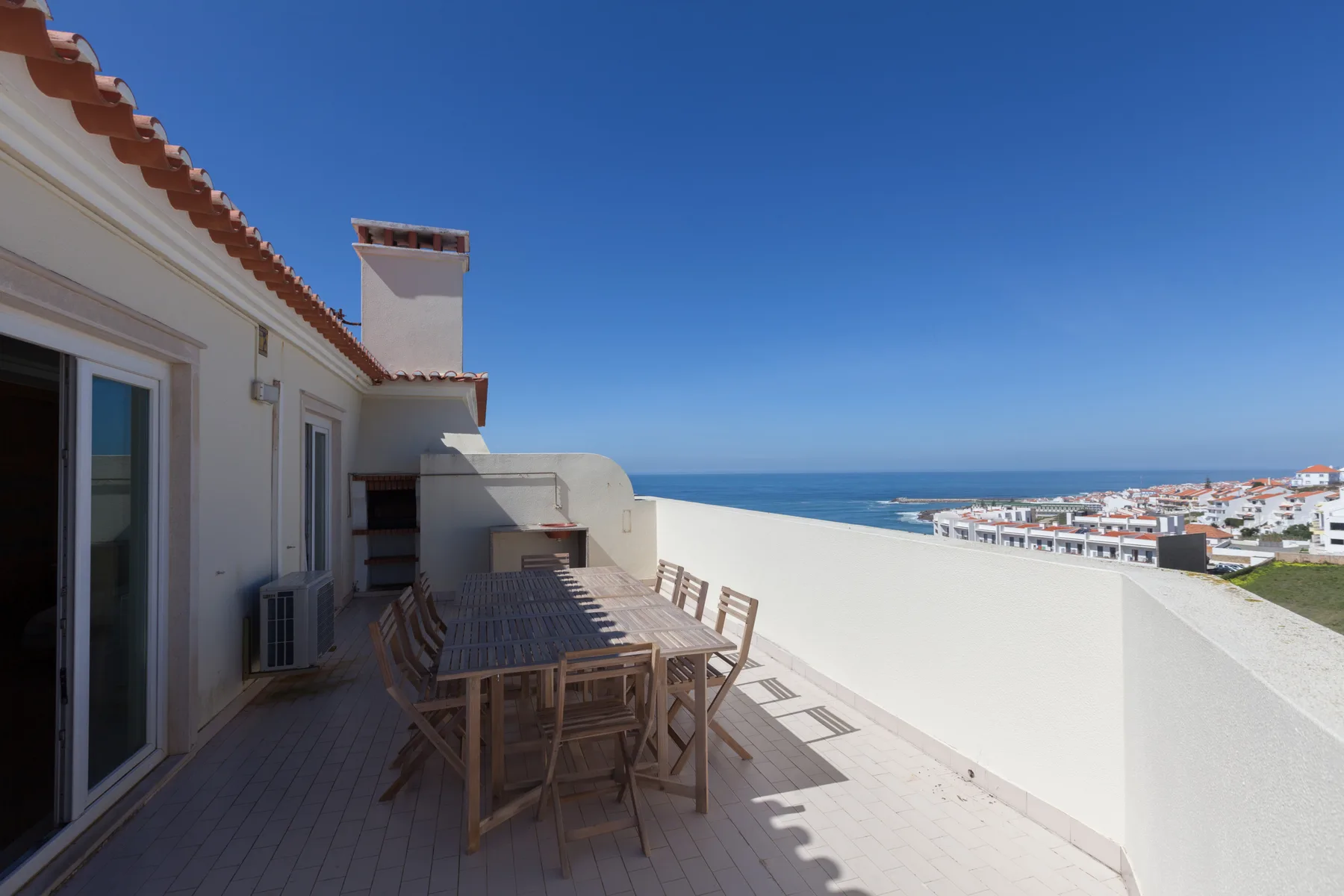 A sunny terrace with outdoor seating overlooks the sea and nearby buildings.