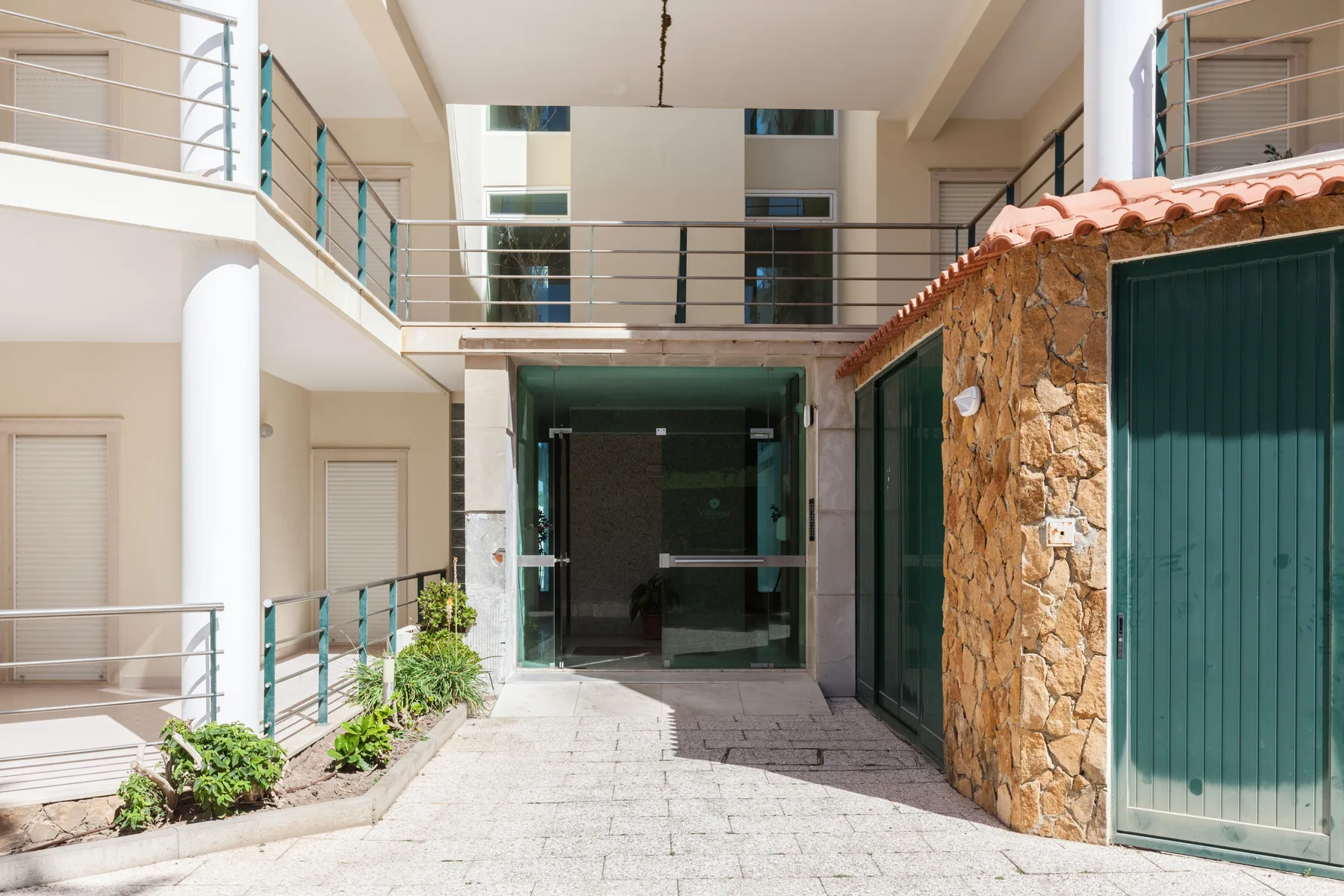 A paved courtyard leads to a glass entrance beside a stone wall and green doors.