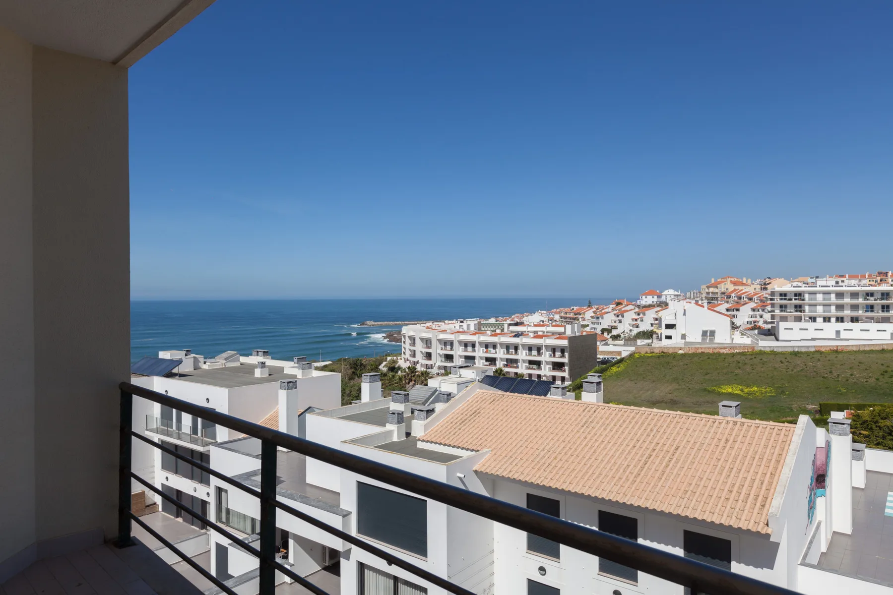 A balcony overlooks coastal buildings and the sea beneath a clear blue sky.