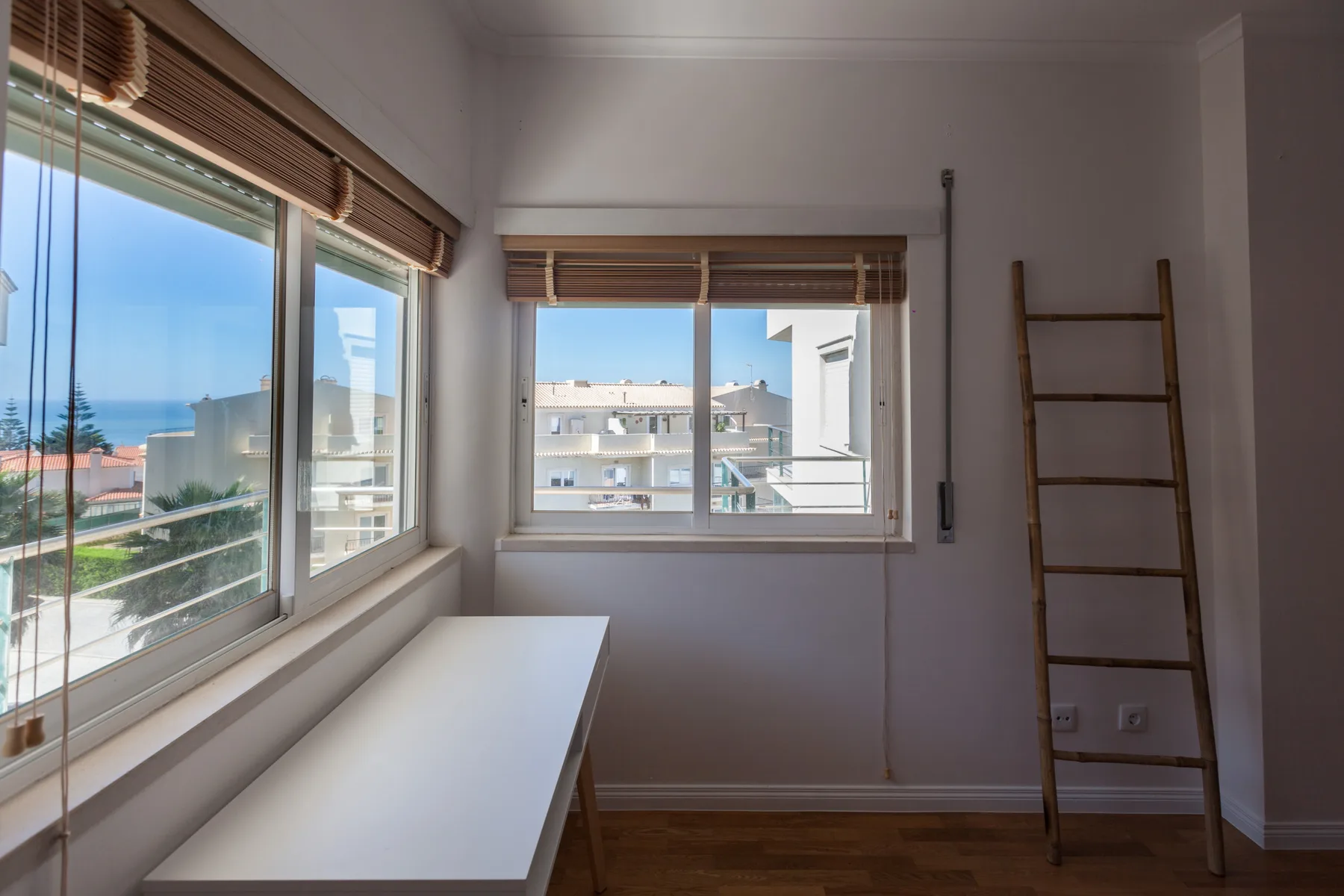 A bright room with wide windows overlooking nearby buildings and the sea.