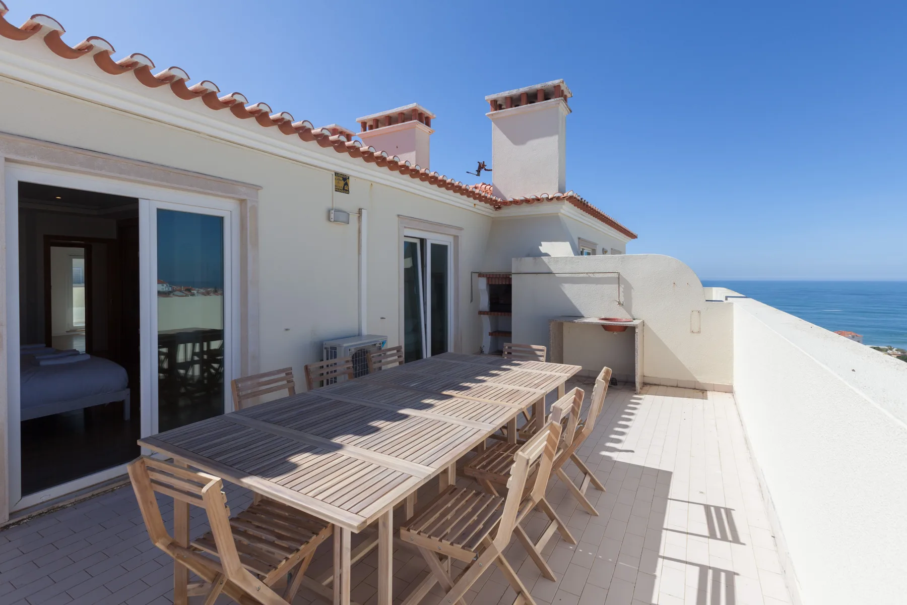 A sunny terrace with outdoor seating and a long dining table beside open glass doors.