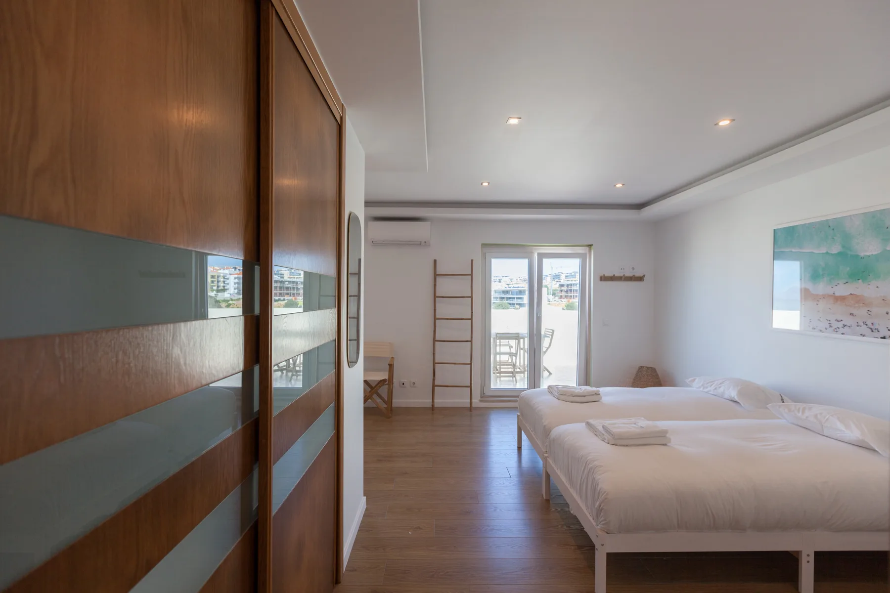 A bright bedroom with beds, wooden flooring, and glass doors opening onto a balcony.