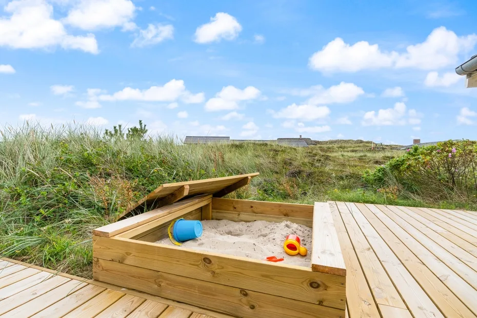 Una zona de juegos de madera al aire libre con arenero está junto a hierbas altas.