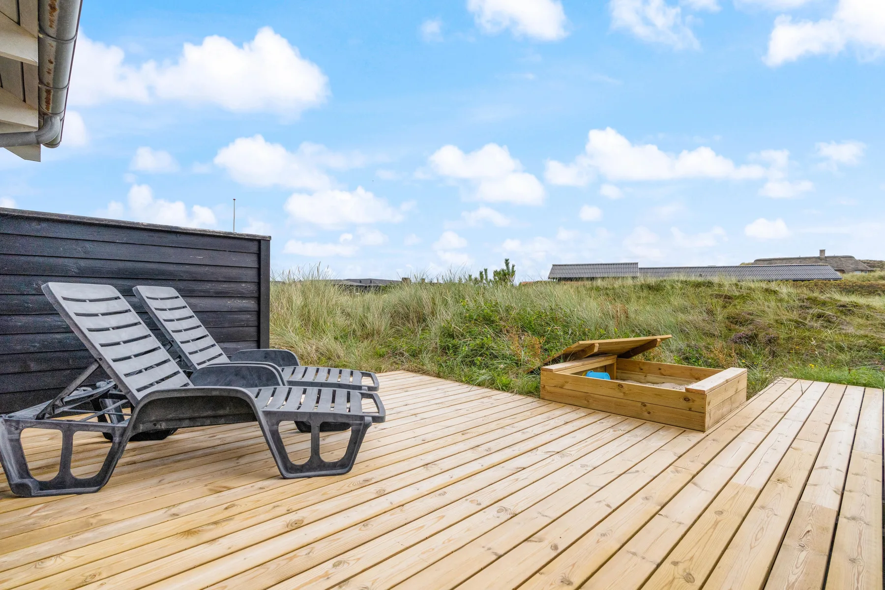 Una terraza de madera con asientos reclinables y una jardinera elevada da a dunas cubiertas de hierba.