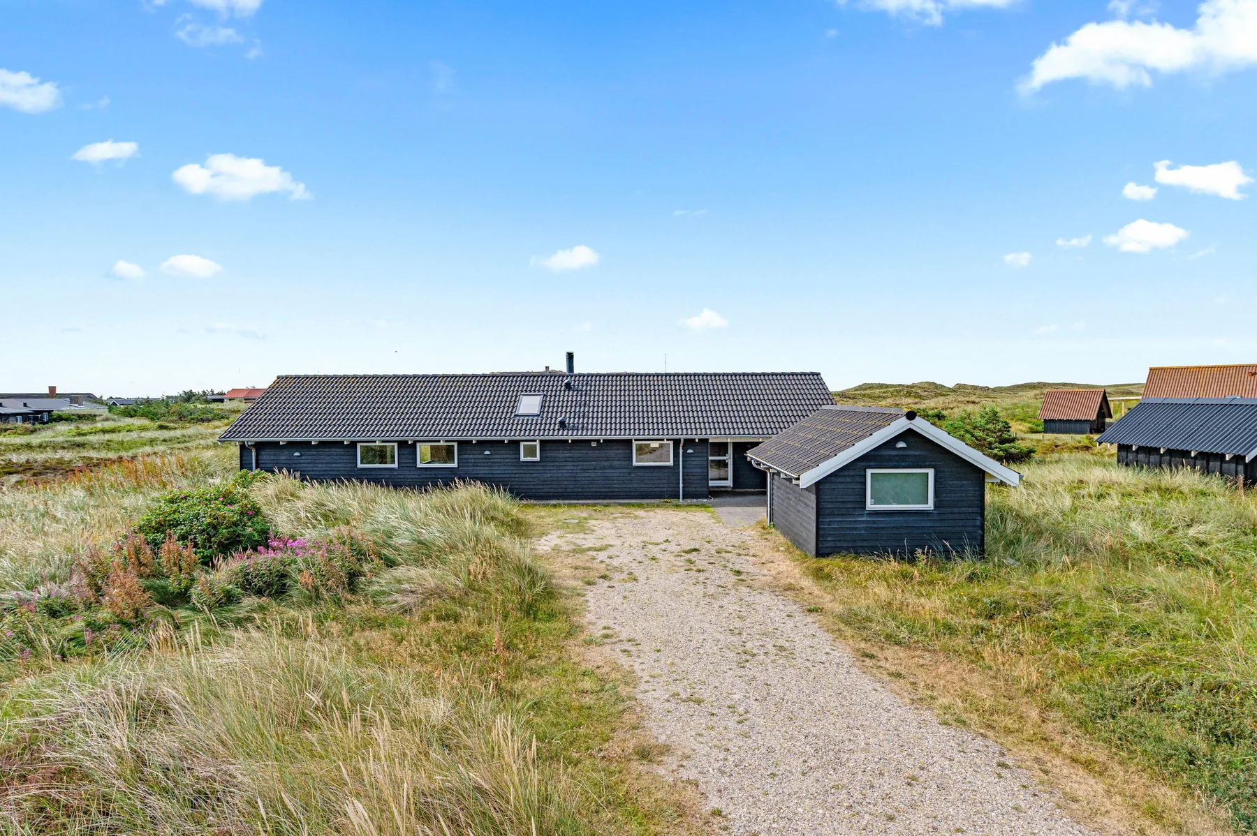 Una casa oscura y baja está junto a un camino de arena entre dunas cubiertas de hierba.