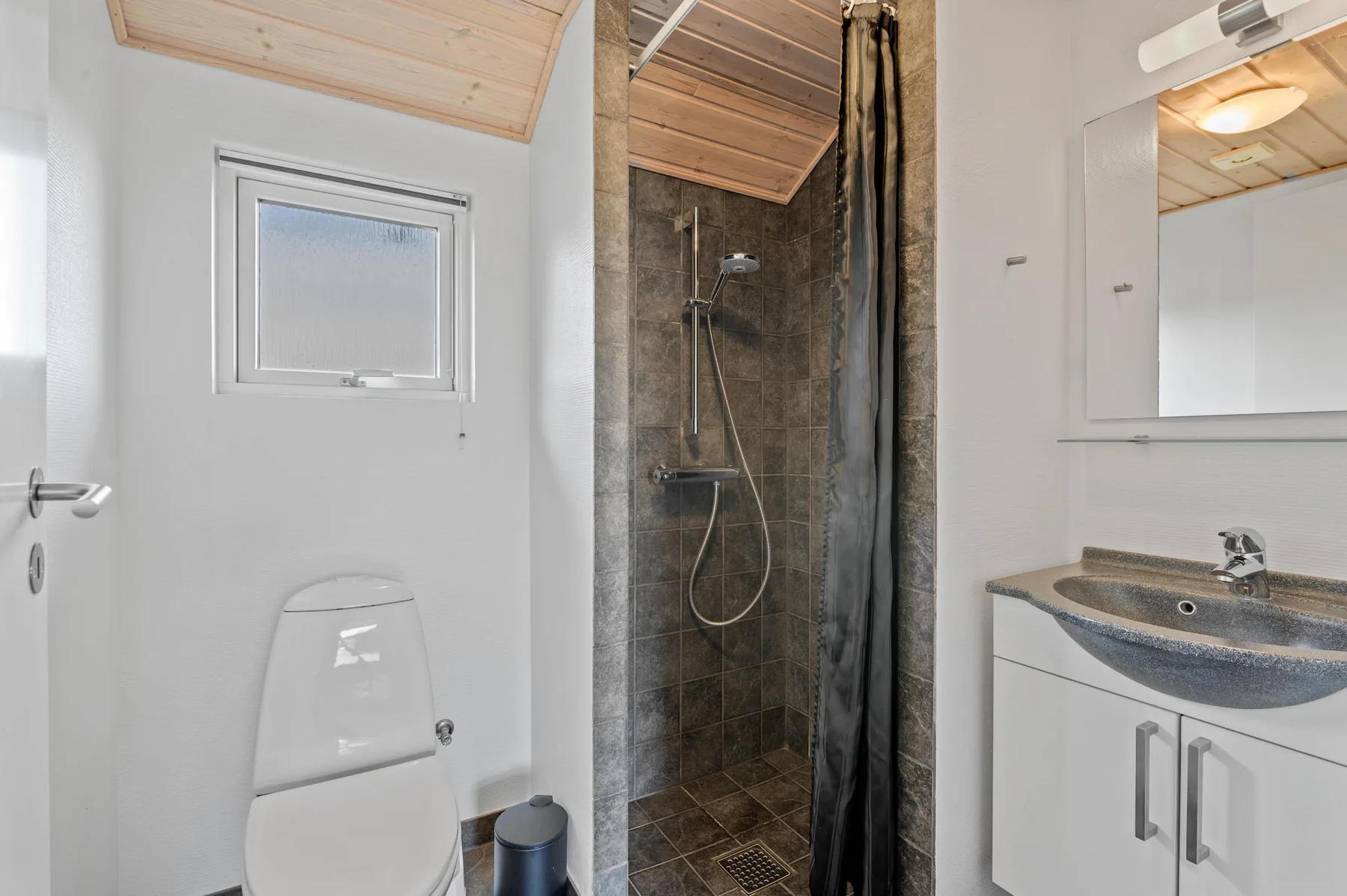Un baño con ducha, inodoro y lavabo bajo un techo de madera inclinado.
