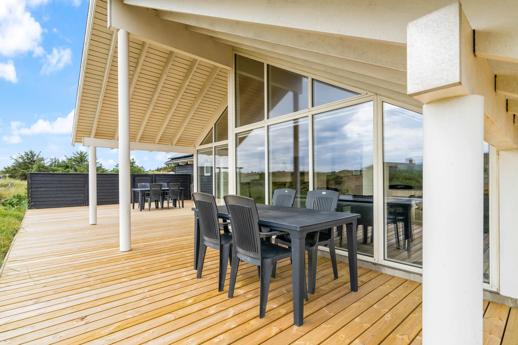 Una terraza de madera cubierta con asientos de comedor al aire libre junto a grandes ventanas.