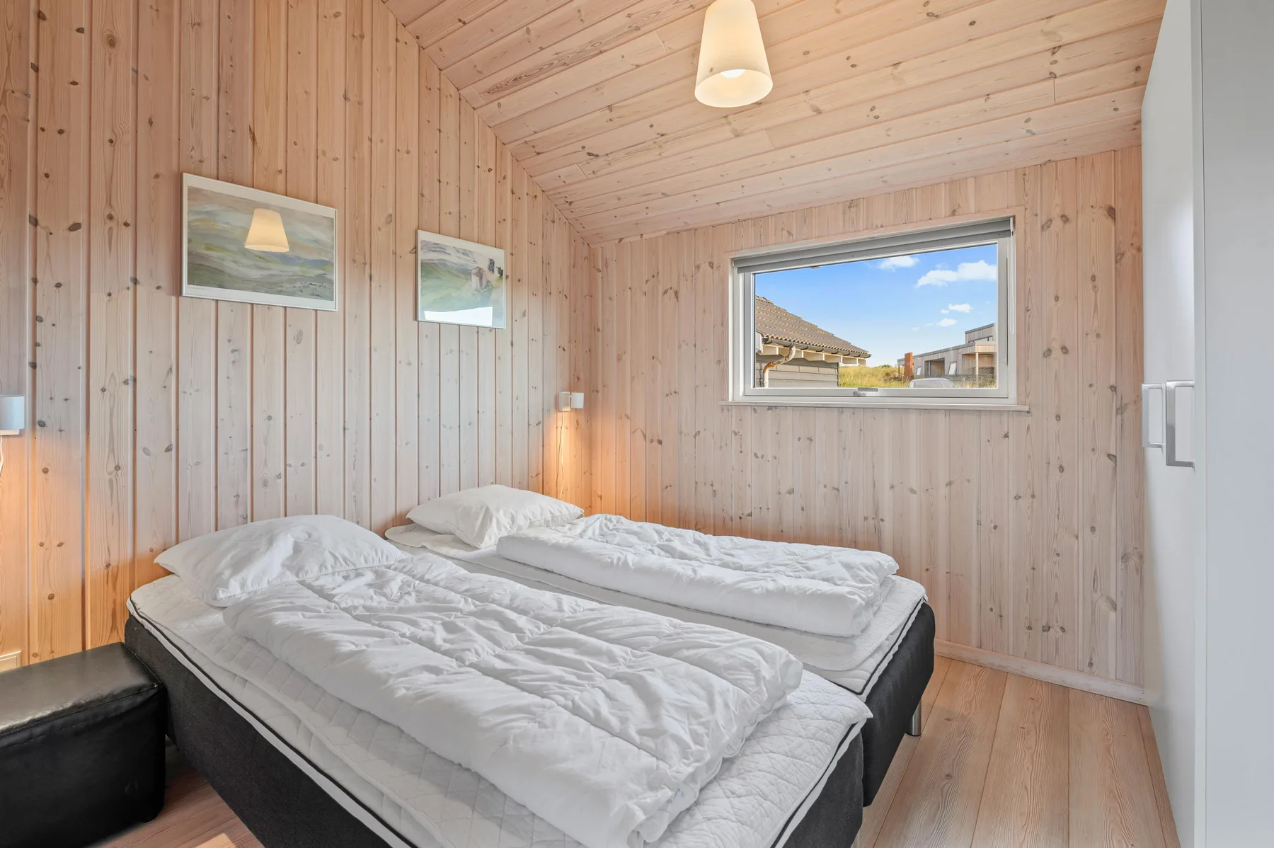 Un dormitorio con dos camas, paredes de madera clara y una ventana con vistas a edificios vecinos.