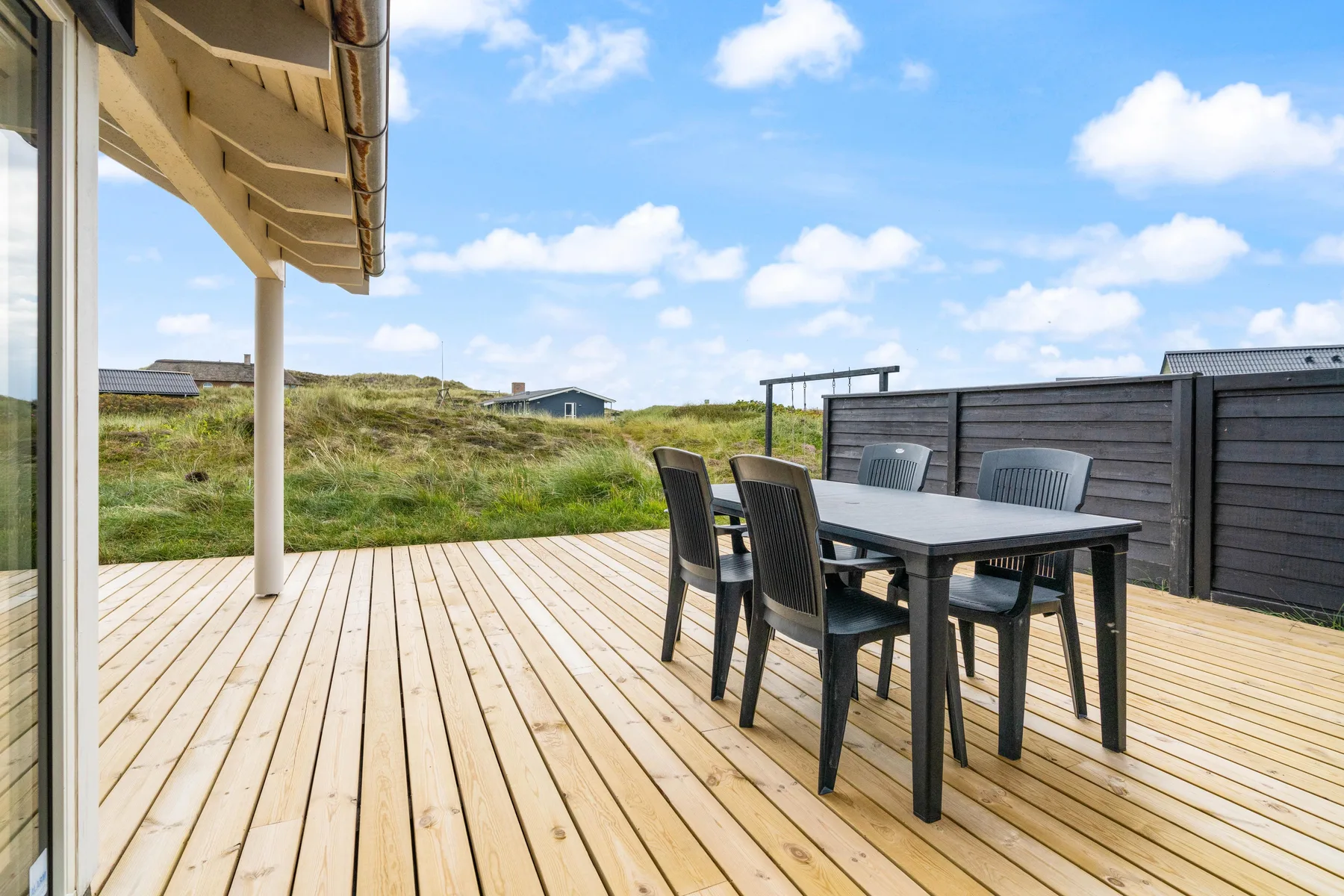 Una terraza de madera tiene una mesa de comedor exterior y asientos junto a un patio cercado.