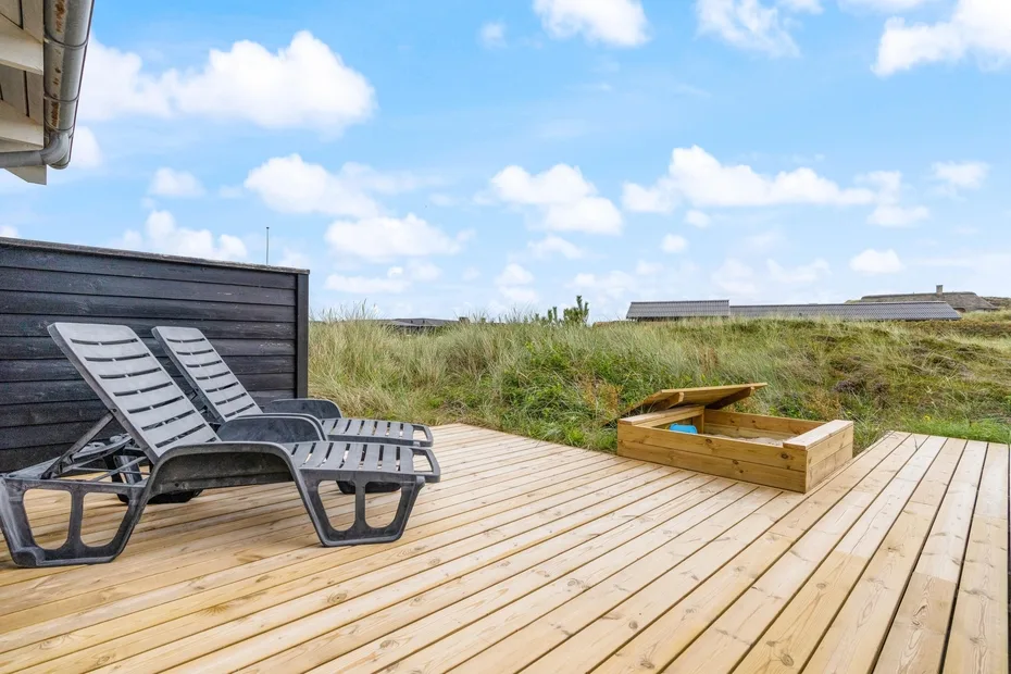 Una terraza exterior de madera con asientos reclinables y una jardinera elevada junto a dunas cubiertas de hierba.