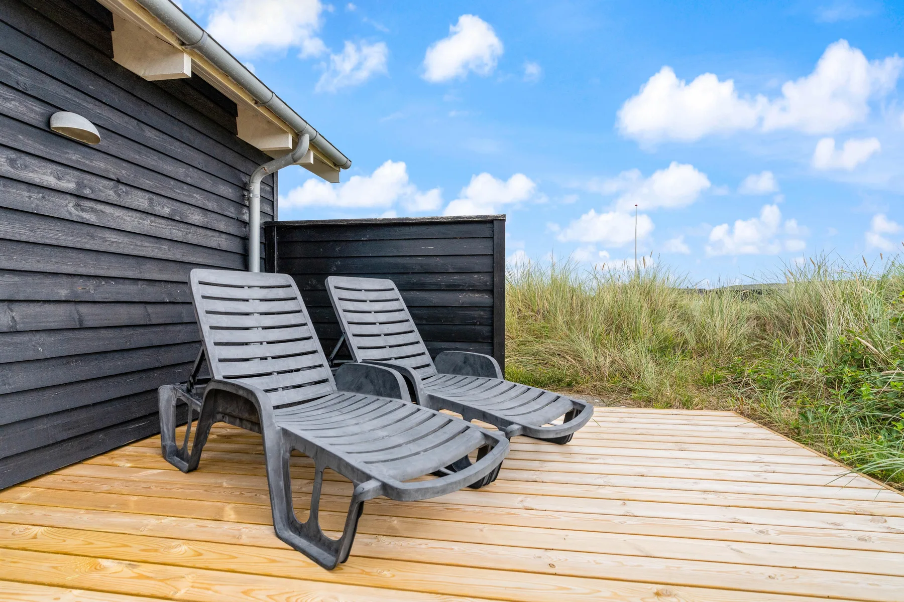 Una terraza de madera junto a una casa oscura tiene sillones reclinables y dunas cubiertas de hierba al fondo.