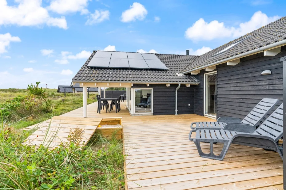 Una terraza de madera junto a una casa de revestimiento oscuro, con asientos exteriores y paneles solares.