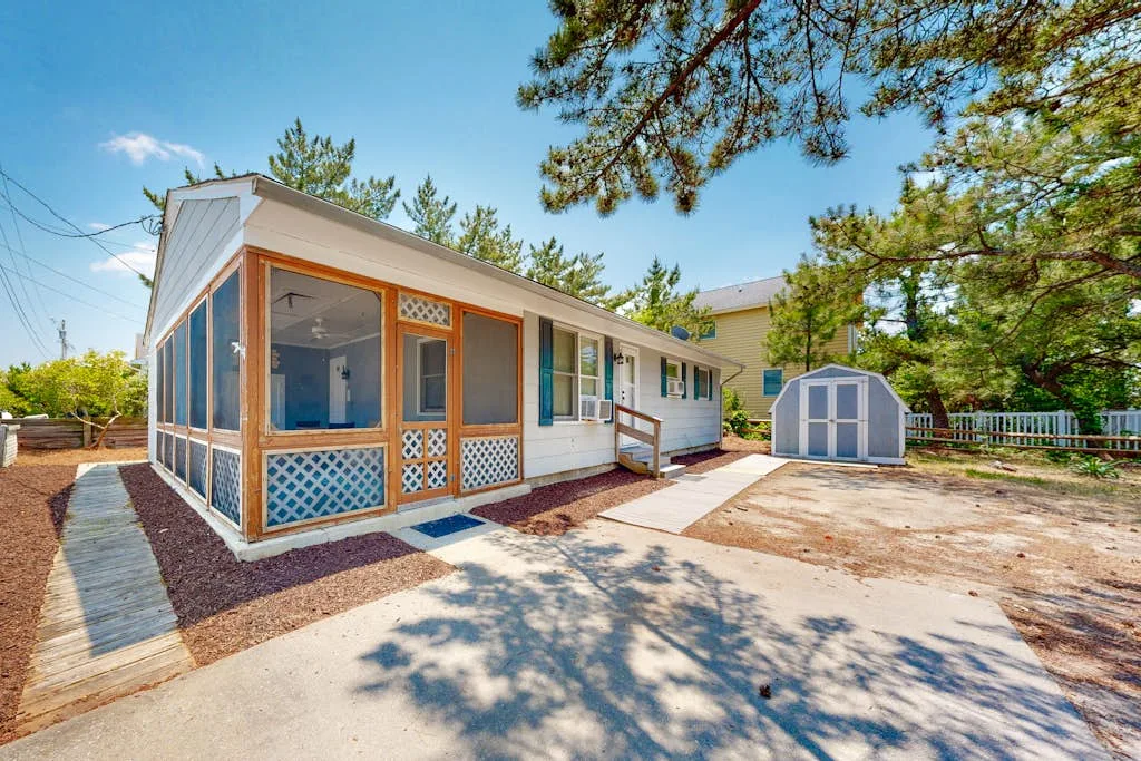 A small house with a screened porch stands beside a shed in a sunny yard.