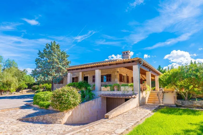 Una casa de piedra con terraza cubierta está junto a un sendero pavimentado y un césped verde.