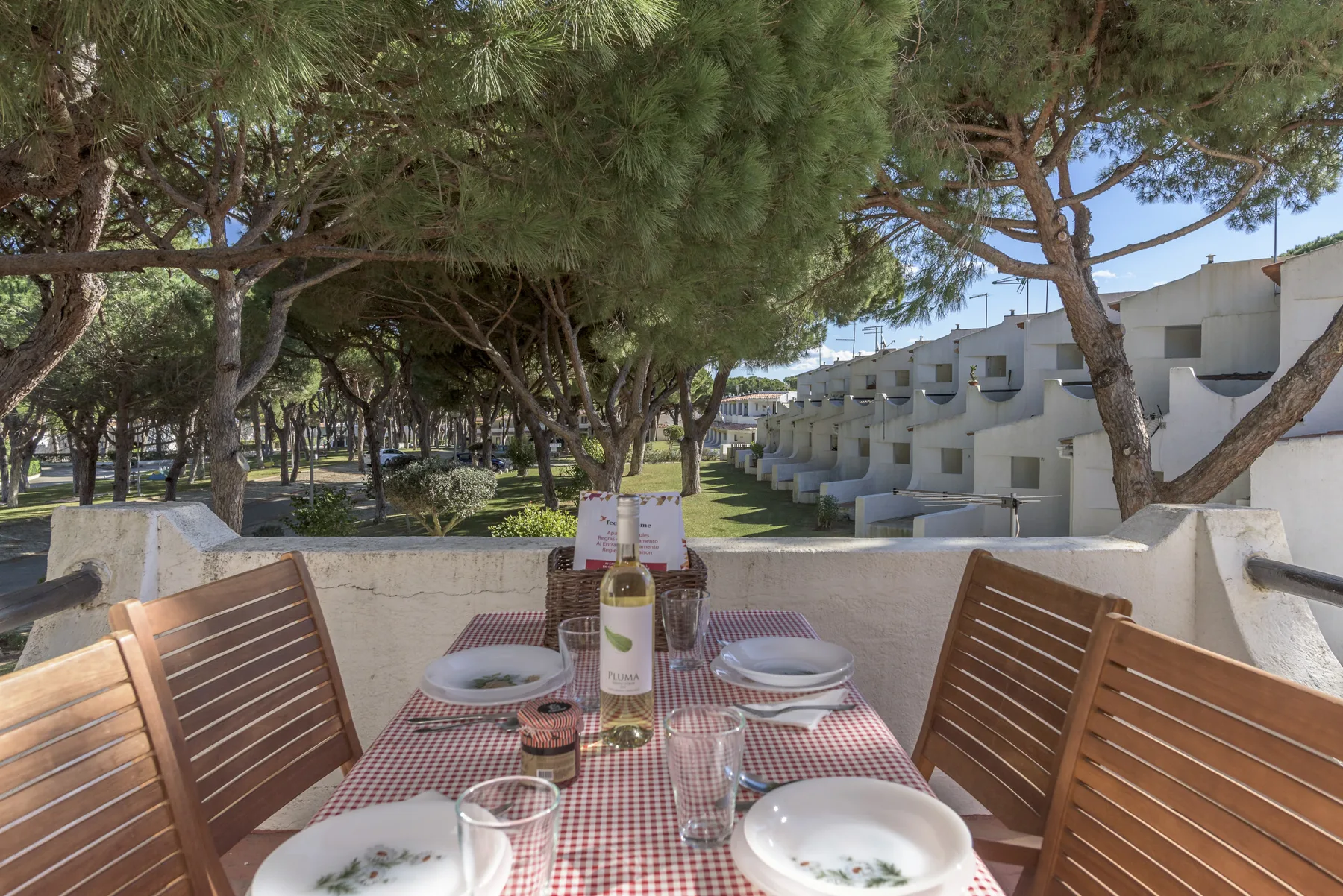 Una mesa al aire libre con cubiertos está bajo los árboles, junto a una fila de edificios.