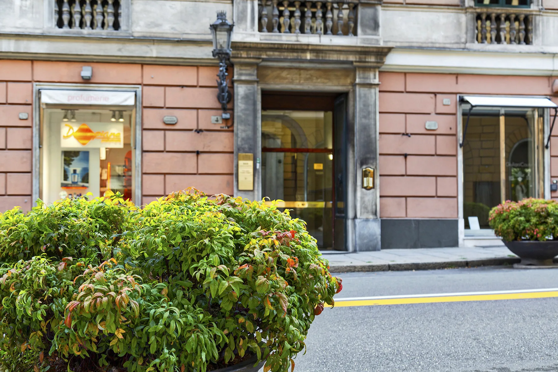 Entrada de un edificio de piedra rosa con grandes ventanas y arbustos frondosos junto a la acera.